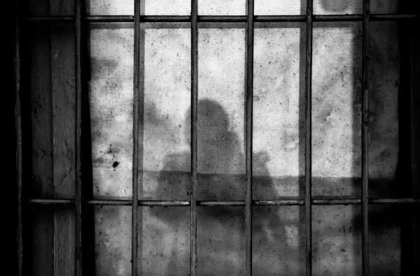 This photo shows a wall, prison bars, and the shadow of a person. It is in black and white.